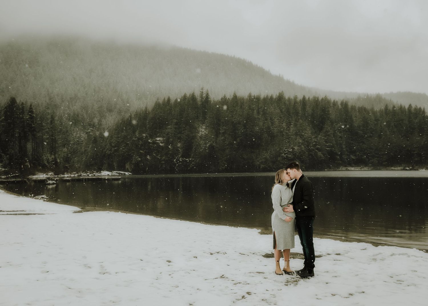 A couple embraces on a snowy shoreline beside a tranquil mountain lake surrounded by evergreen forest.
