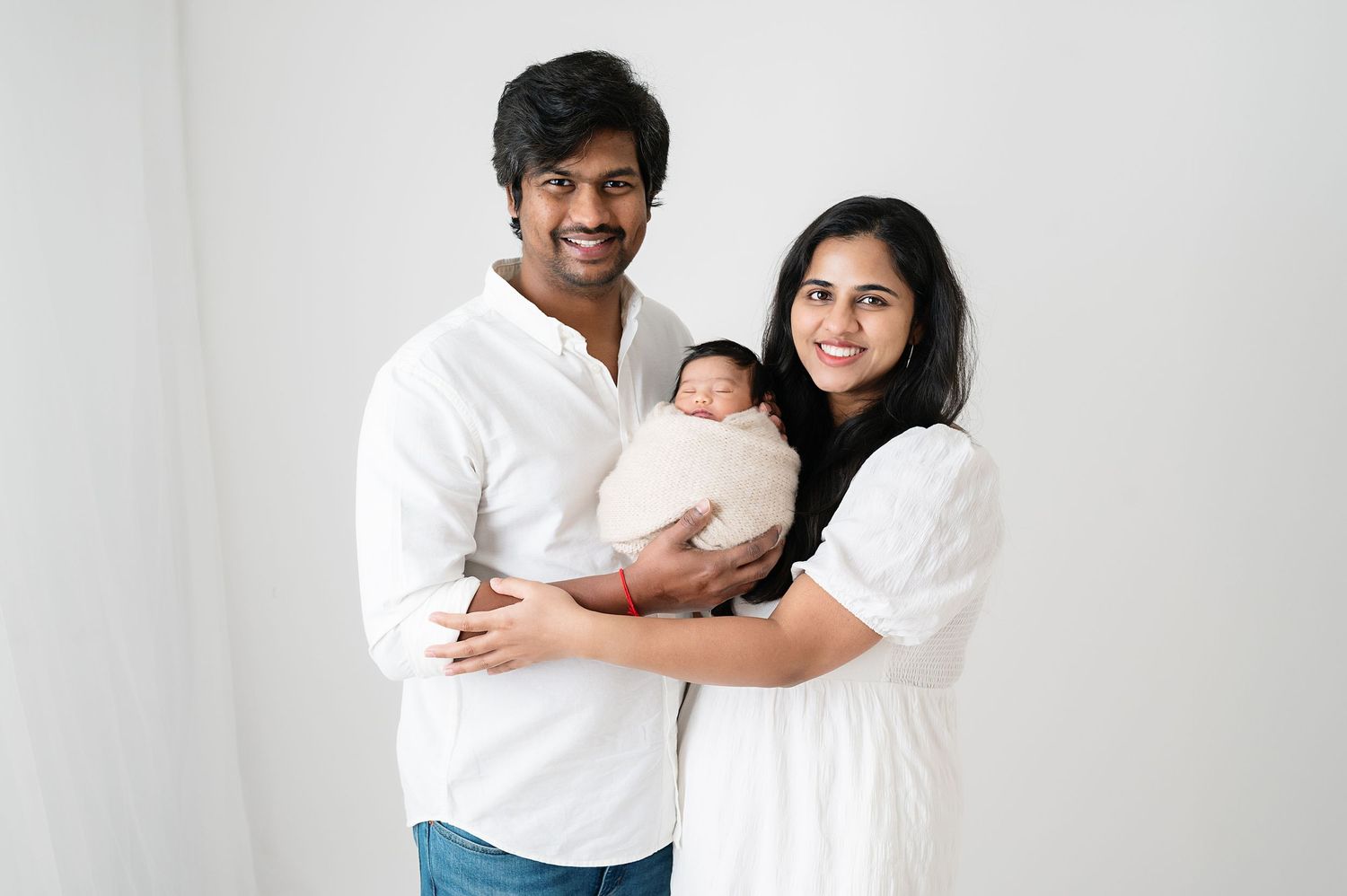 Parents in white outfits holding newborn against clean white background during studio photography session.
