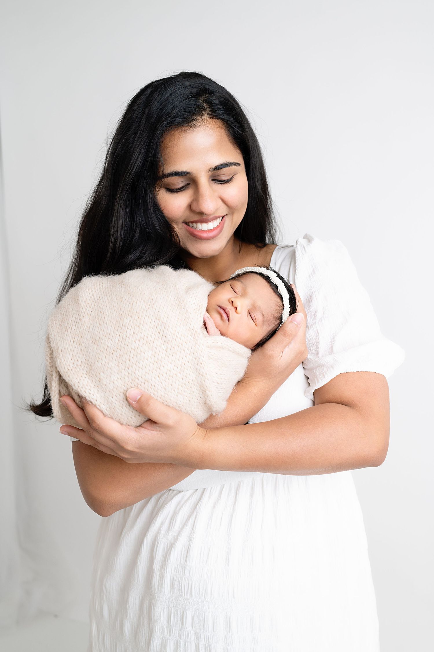 Mother in white dress holding swaddled infant against clean white studio backdrop.