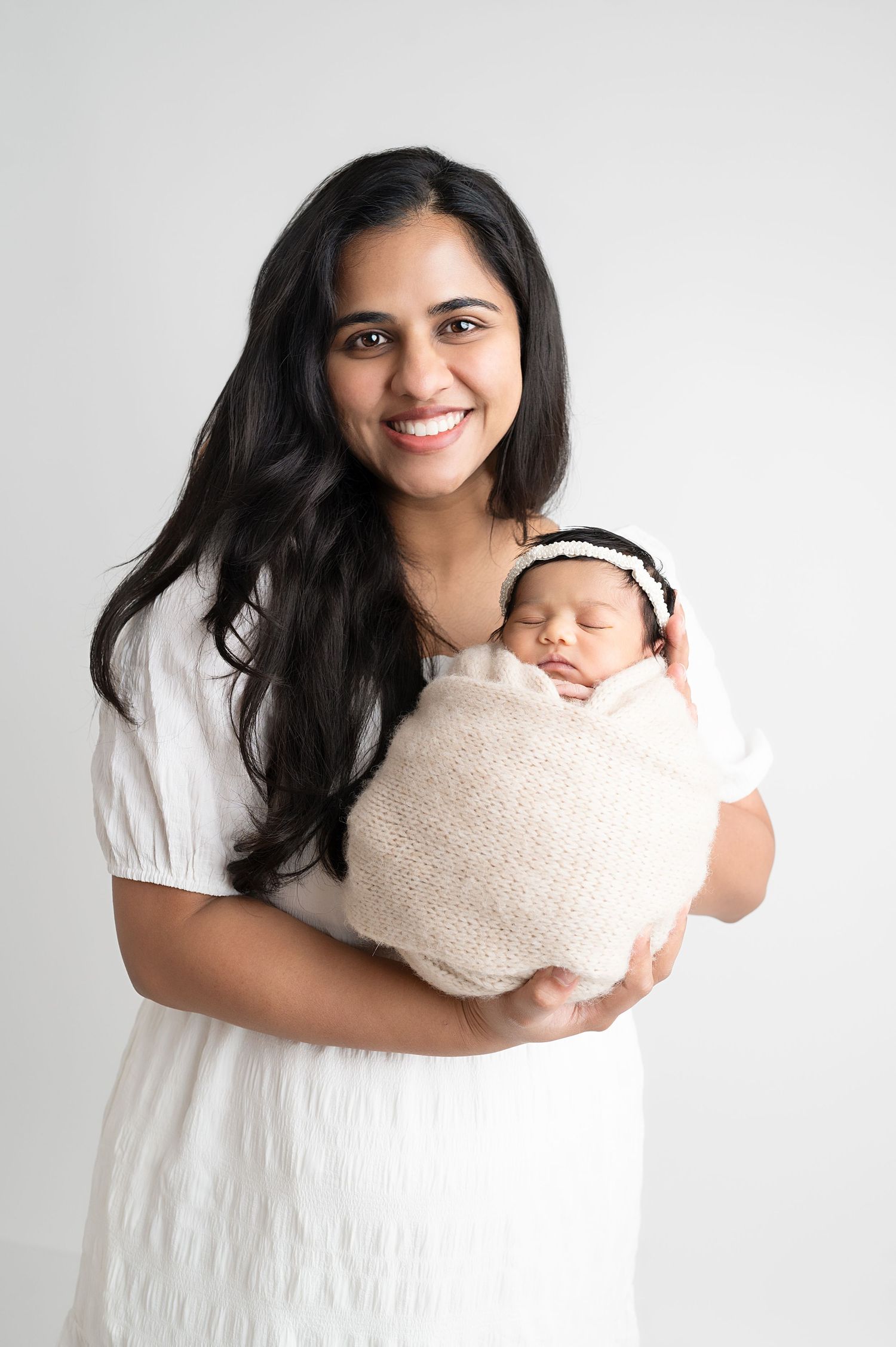 Studio portraits of mother in white dress holding newborn wrapped in beige blanket.