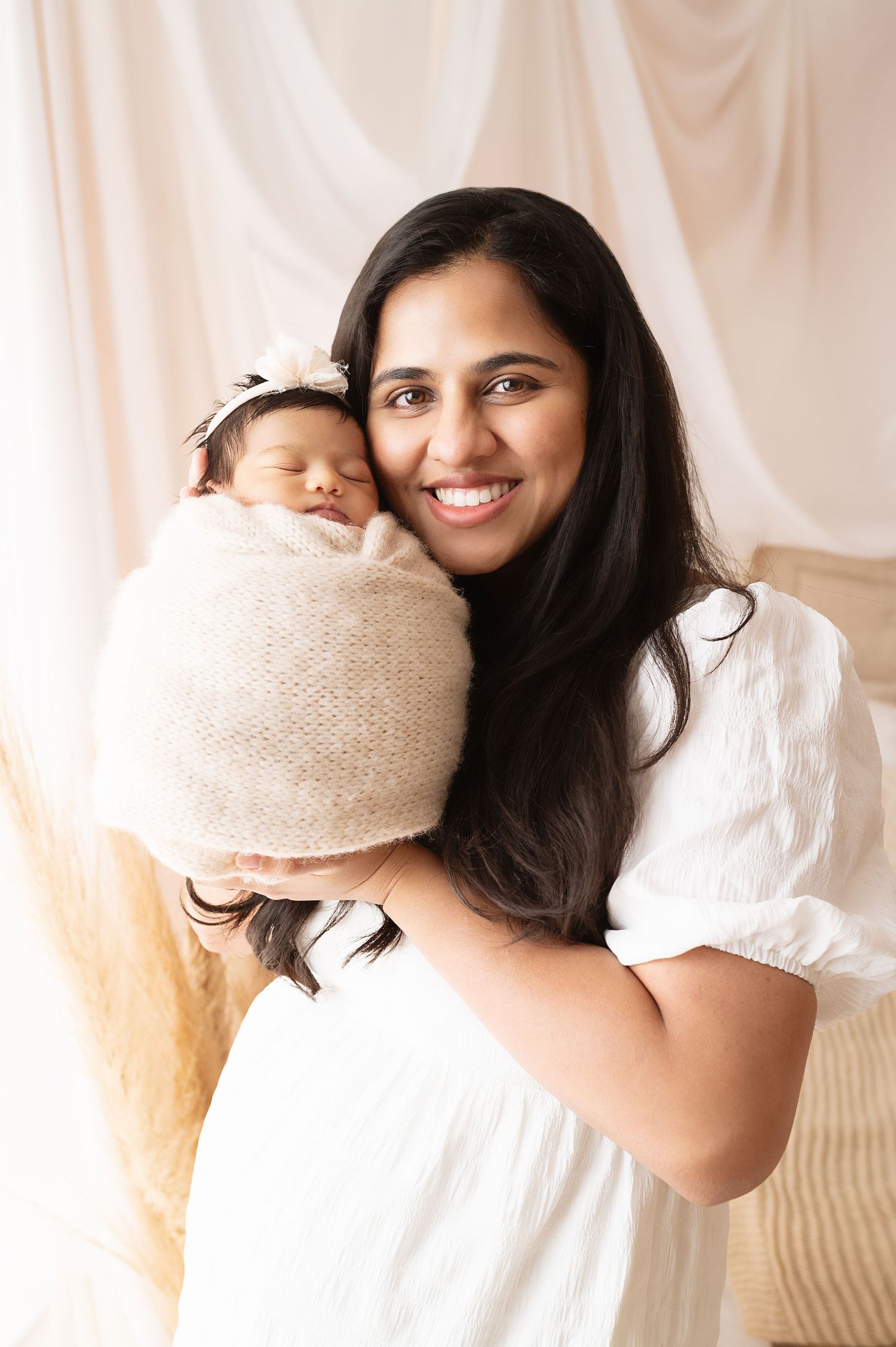 Parent in white dress snuggling newborn wrapped in beige knit blanket.