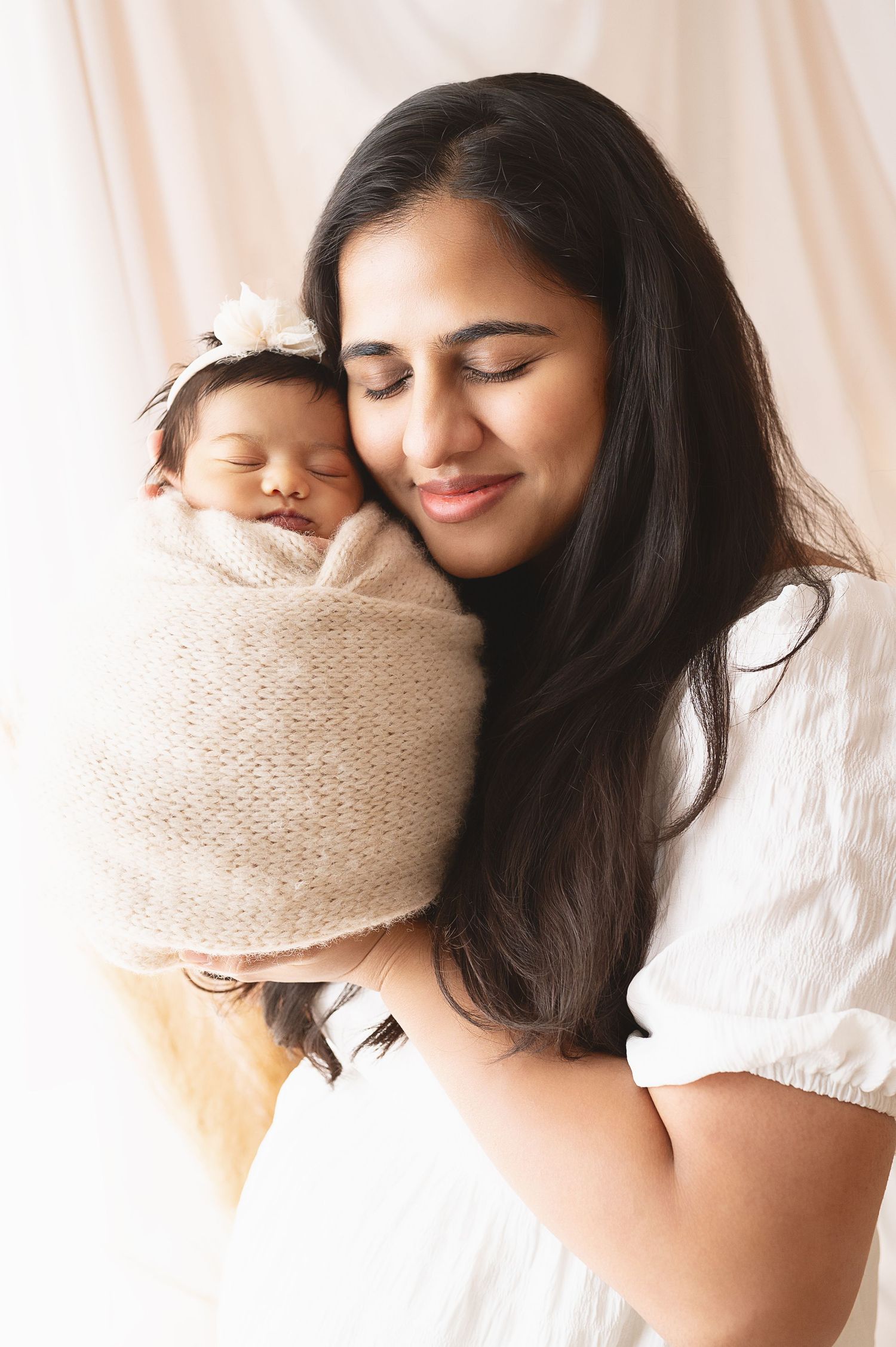 Loving parent in white dress holding sleeping infant wrapped in beige knit blanket.