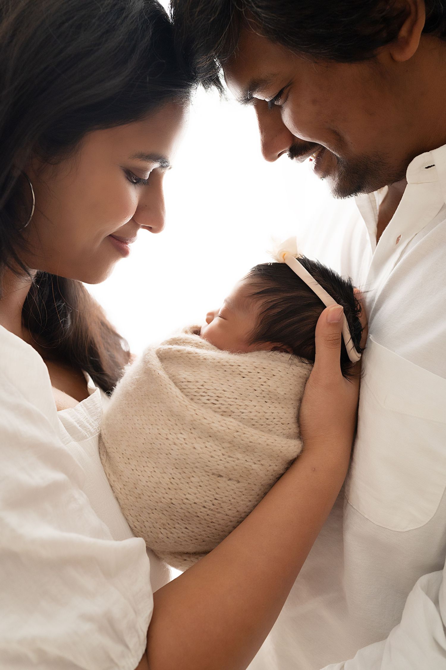 Parents in white sharing intimate moment with their swaddled newborn.