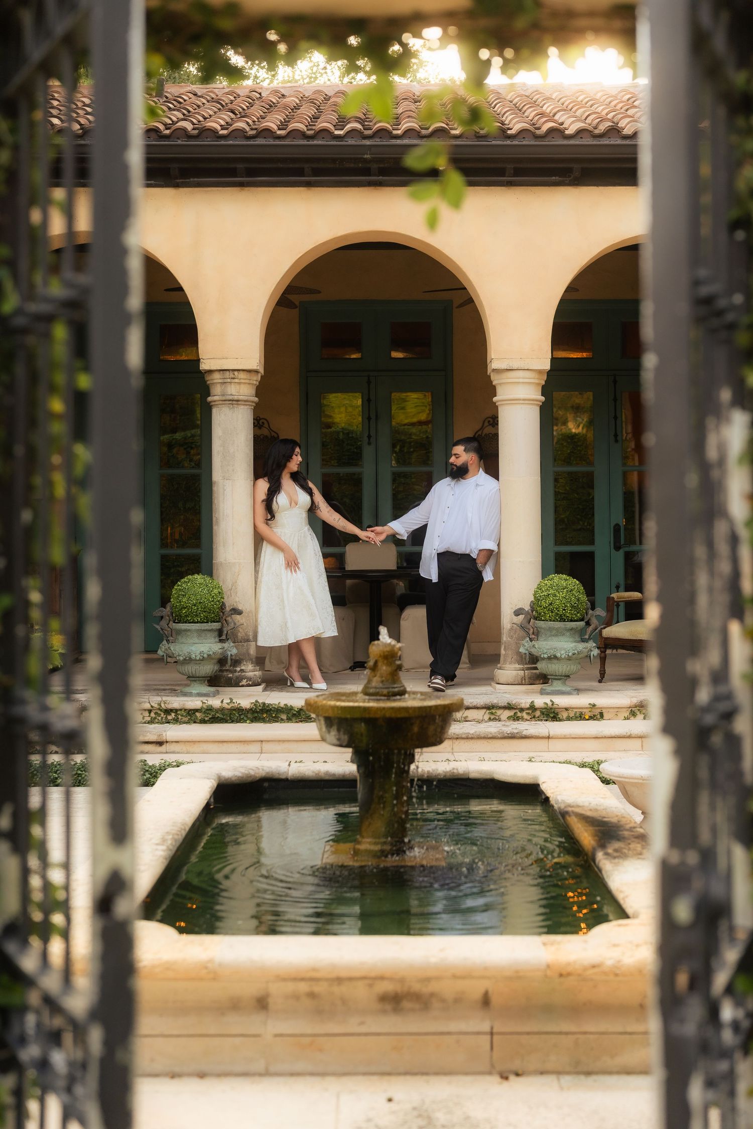 Engaged couple holding hands by a fountain in an elegant Mediterranean-style courtyard.