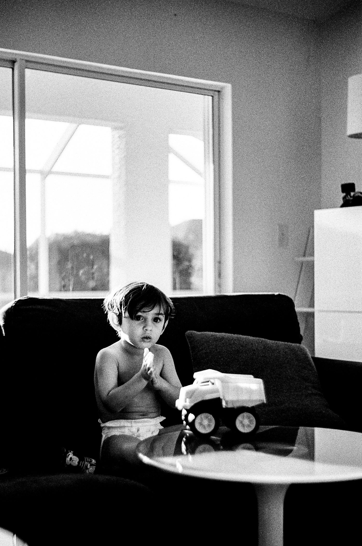 Black and white photo of a child sitting on a couch near a window with a toy truck visible on the table.
