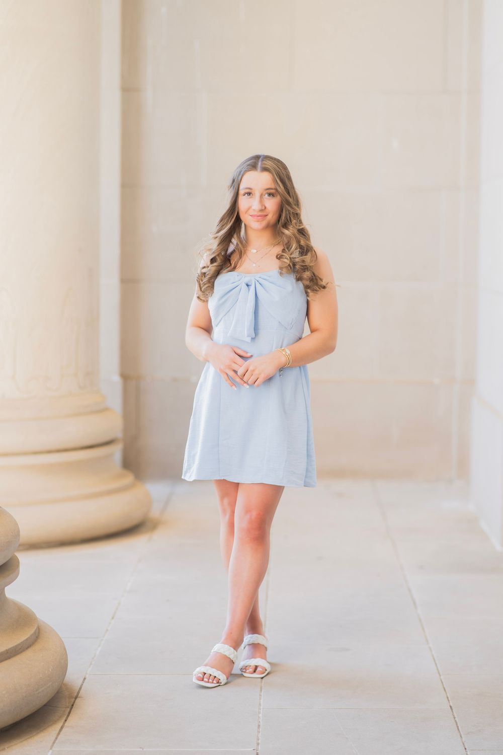 Teen girl in light blue bow-front sundress poses between stone columns for senior portraits.