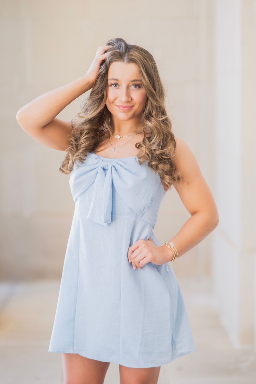 Smiling teen girl in light blue bow-front dress poses with hand in hair for senior photos.