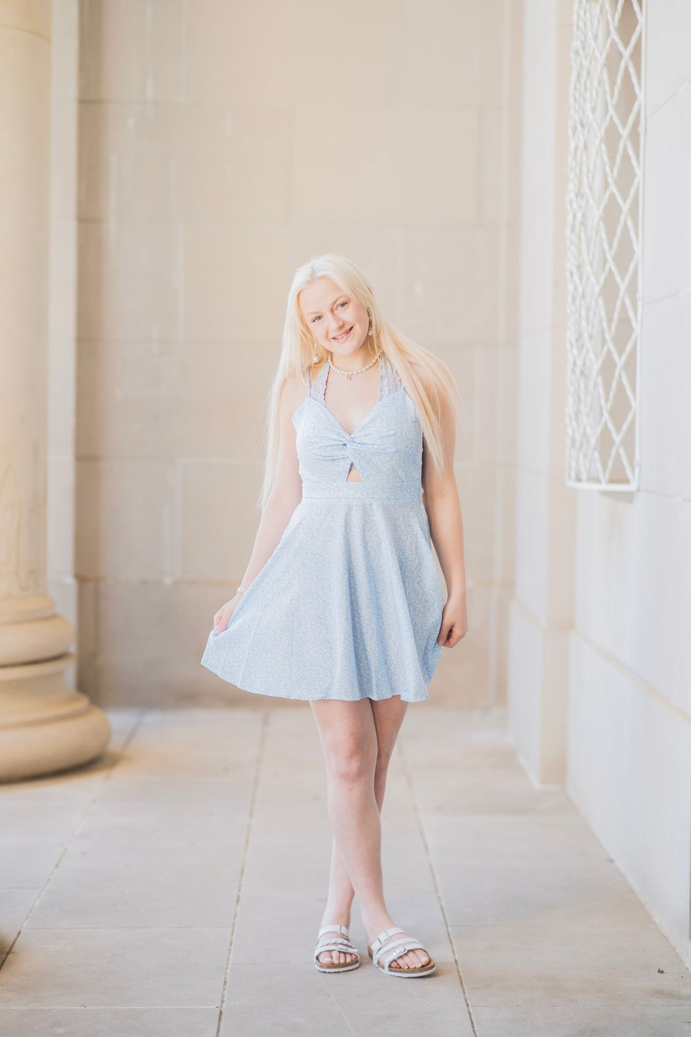 Young woman with long blonde hair smiling in a light blue halter mini dress and white sandals outdoors.