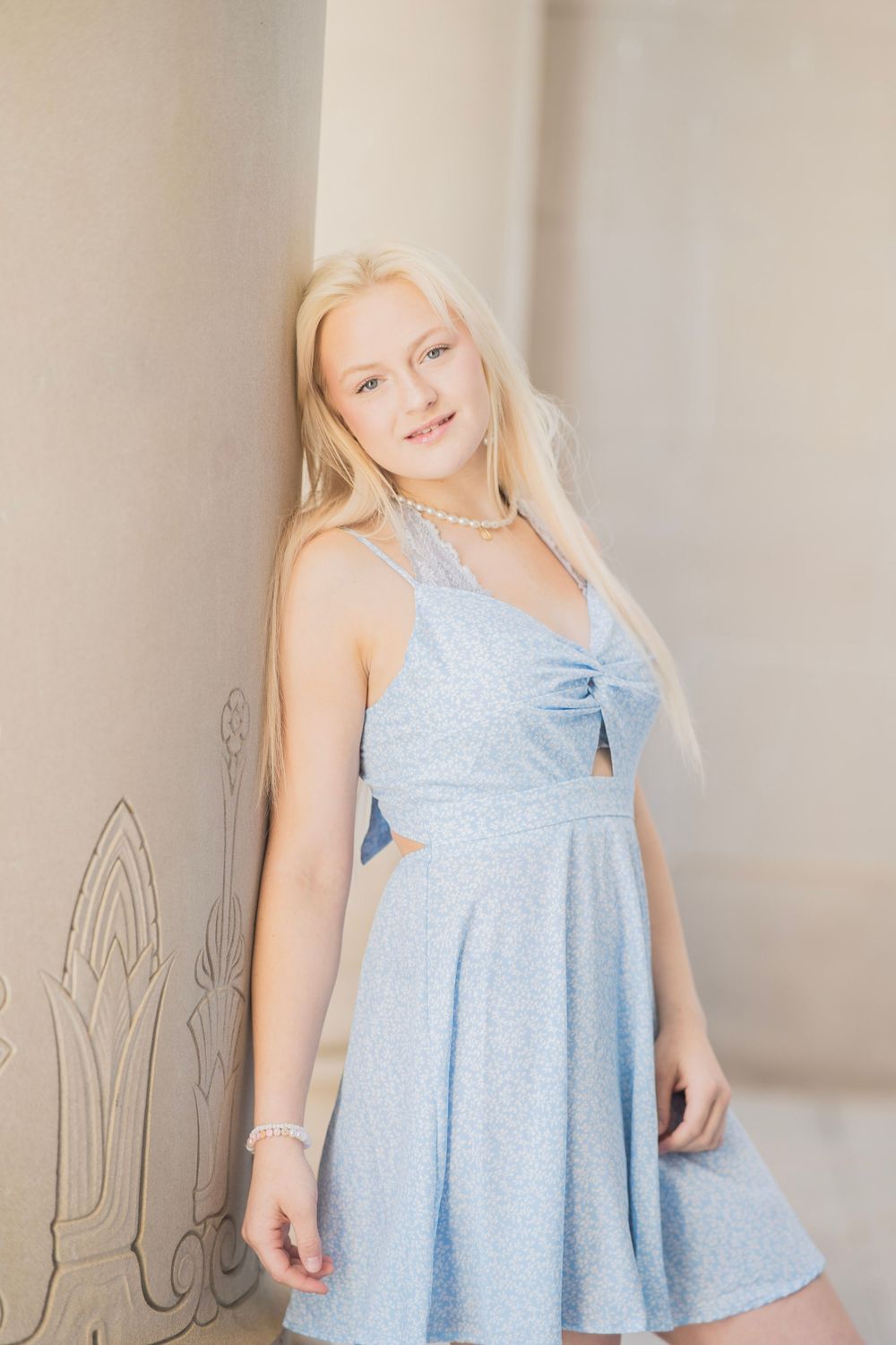 Blonde teenage girl in a light blue sleeveless dress leaning against a stone column wearing pearl jewelry.