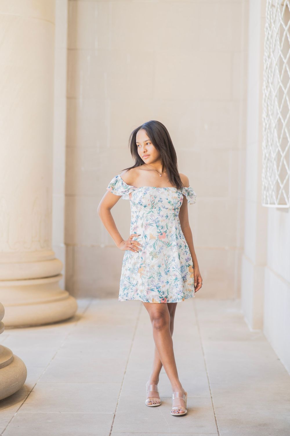 Young woman in off-shoulder floral mini dress poses between stone columns in natural light.