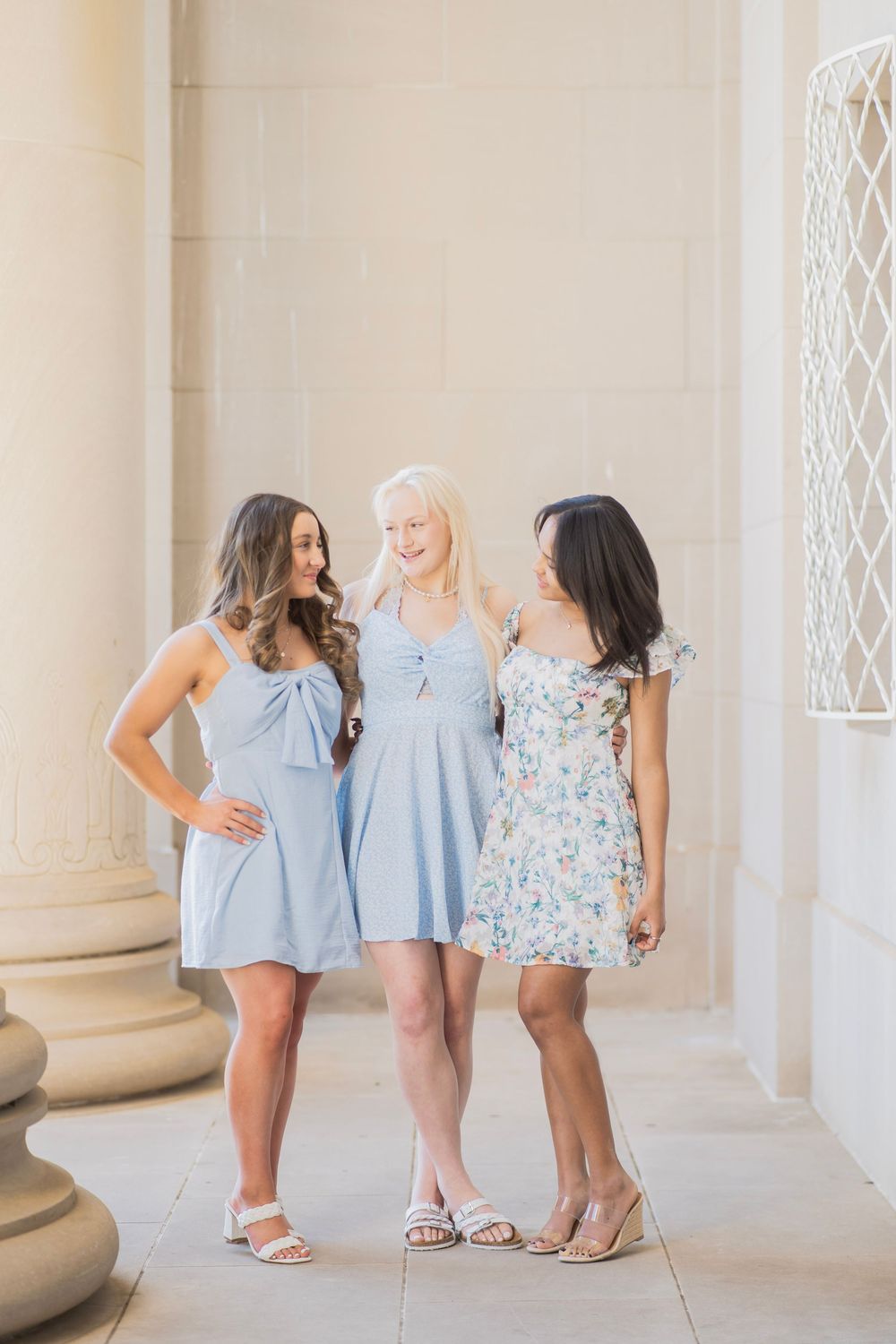 Three young women in pastel summer dresses posing together between stone columns in bright sunlight.