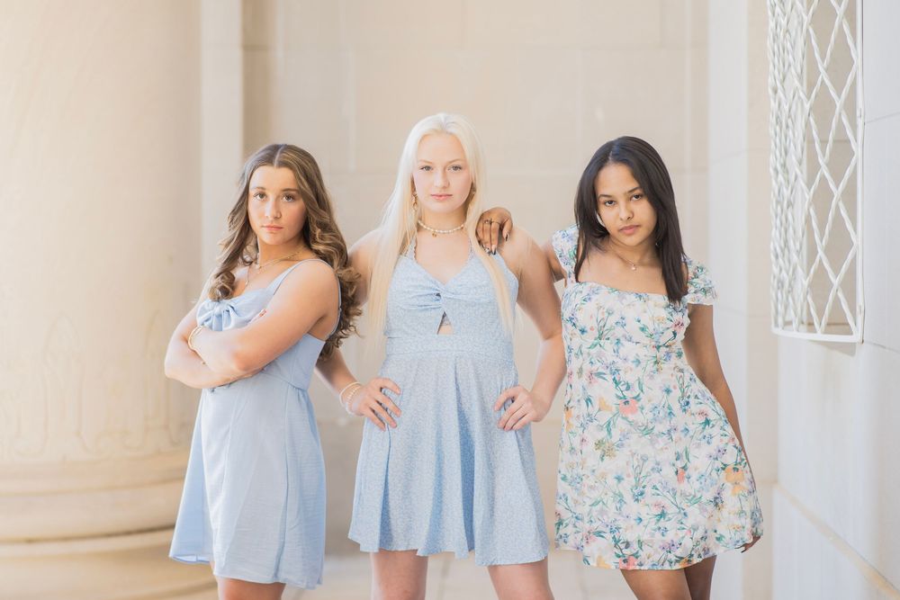 Three teenage girls in pastel dresses striking a serious pose between white columns in an elegant setting.