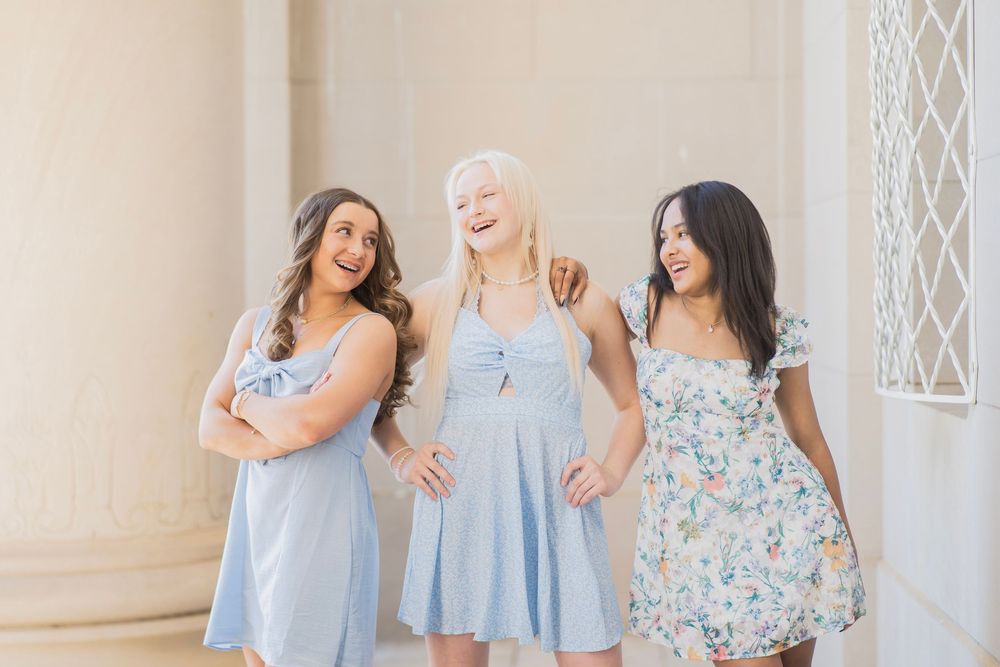 Three laughing young women in light blue and floral summer dresses standing between stone pillars outdoors.