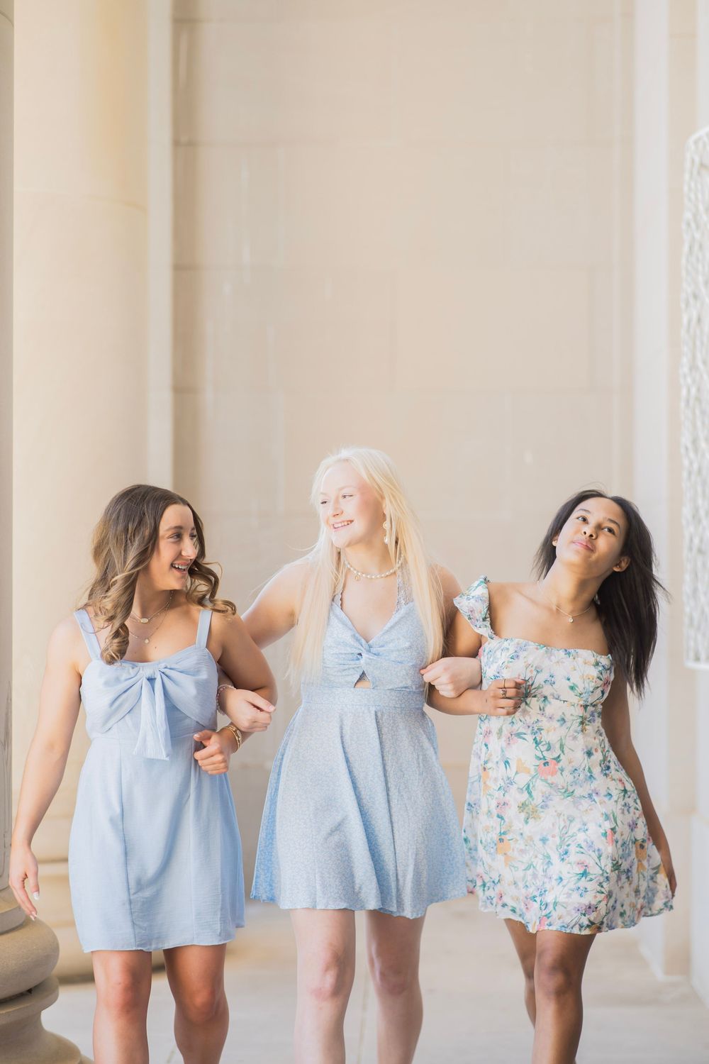 Three smiling young women in pastel dresses walking arm in arm between elegant stone columns outdoors.