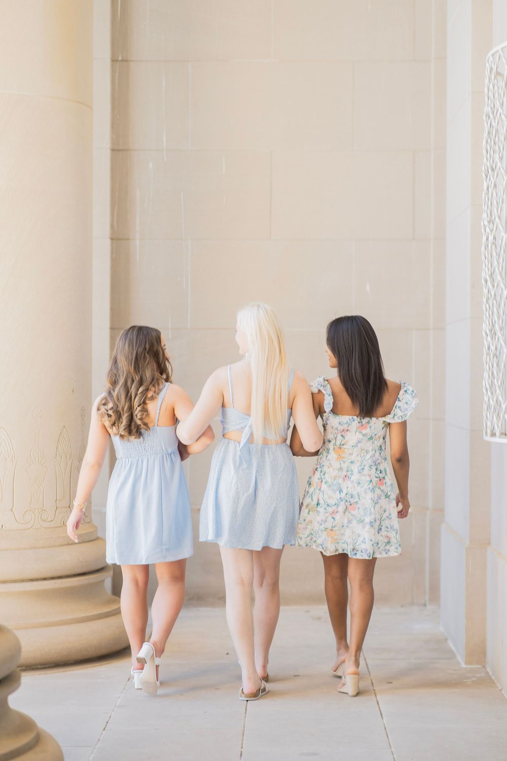 Back view of three young women in pastel summer dresses walking arm in arm between stone columns.