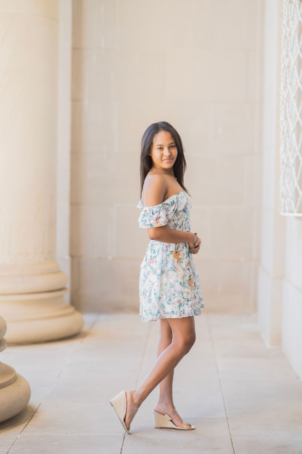 Teen girl in floral off-shoulder dress poses gracefully between stone columns in warm sunlight.