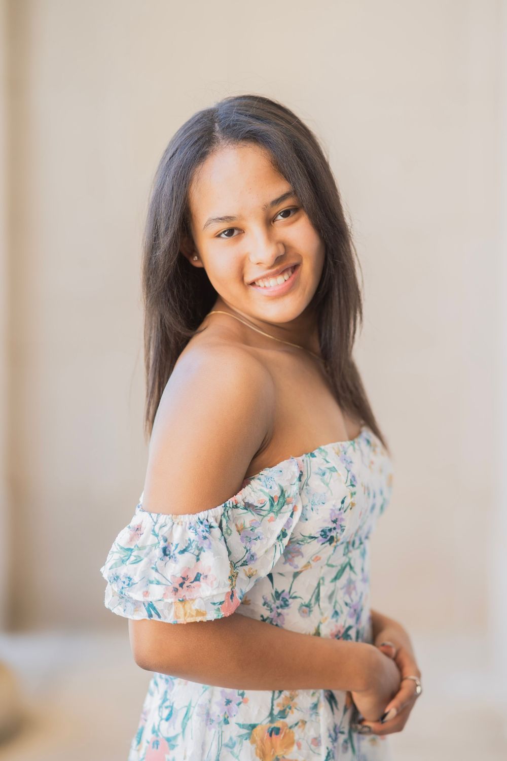 Smiling teen girl in off-shoulder floral dress poses for senior portrait in warm natural light.