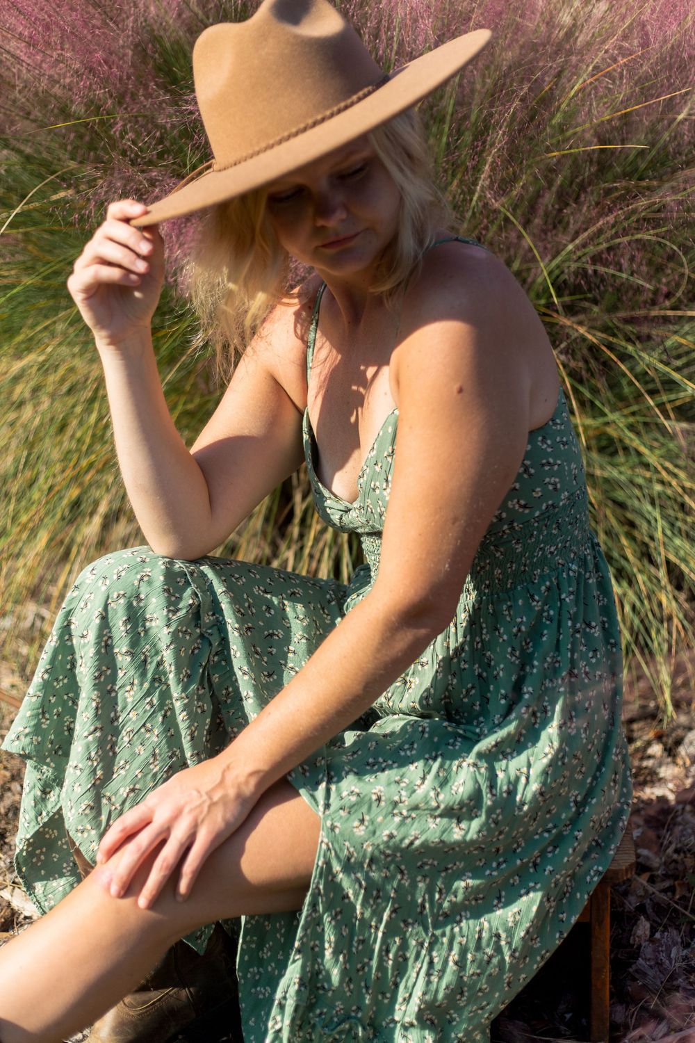 A person in a flowy green floral dress and tan sun hat poses against tall ornamental grasses at sunset.