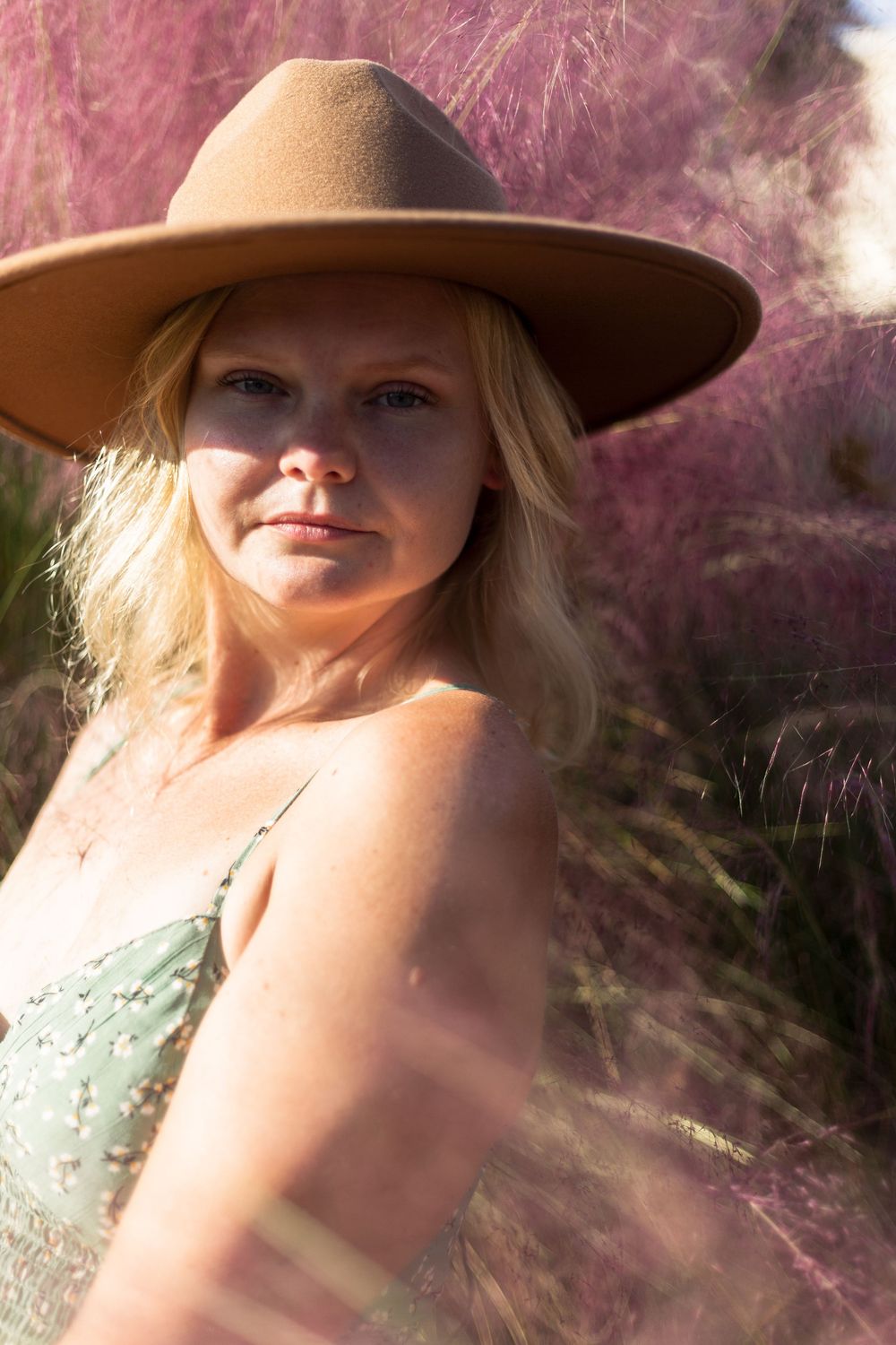 A person in a brown wide-brimmed hat poses outdoors against soft pink foliage wearing a sage green top on a sunny day.