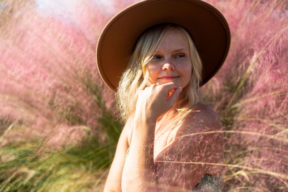 A person in a brown hat poses thoughtfully among pink grass during golden hour.