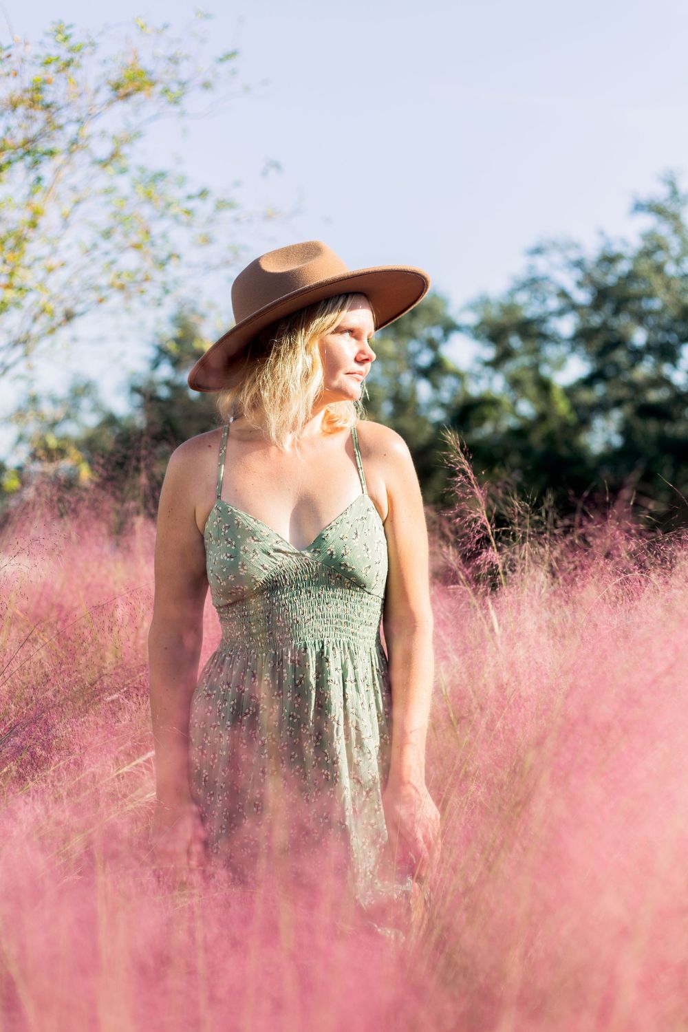 A person in a green sundress and brown hat stands among pink wildflowers on a bright summer day.