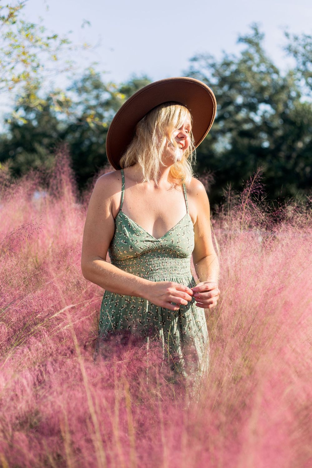 A person in a floral dress and brown hat stands among pink flowering grass in summer sunlight.