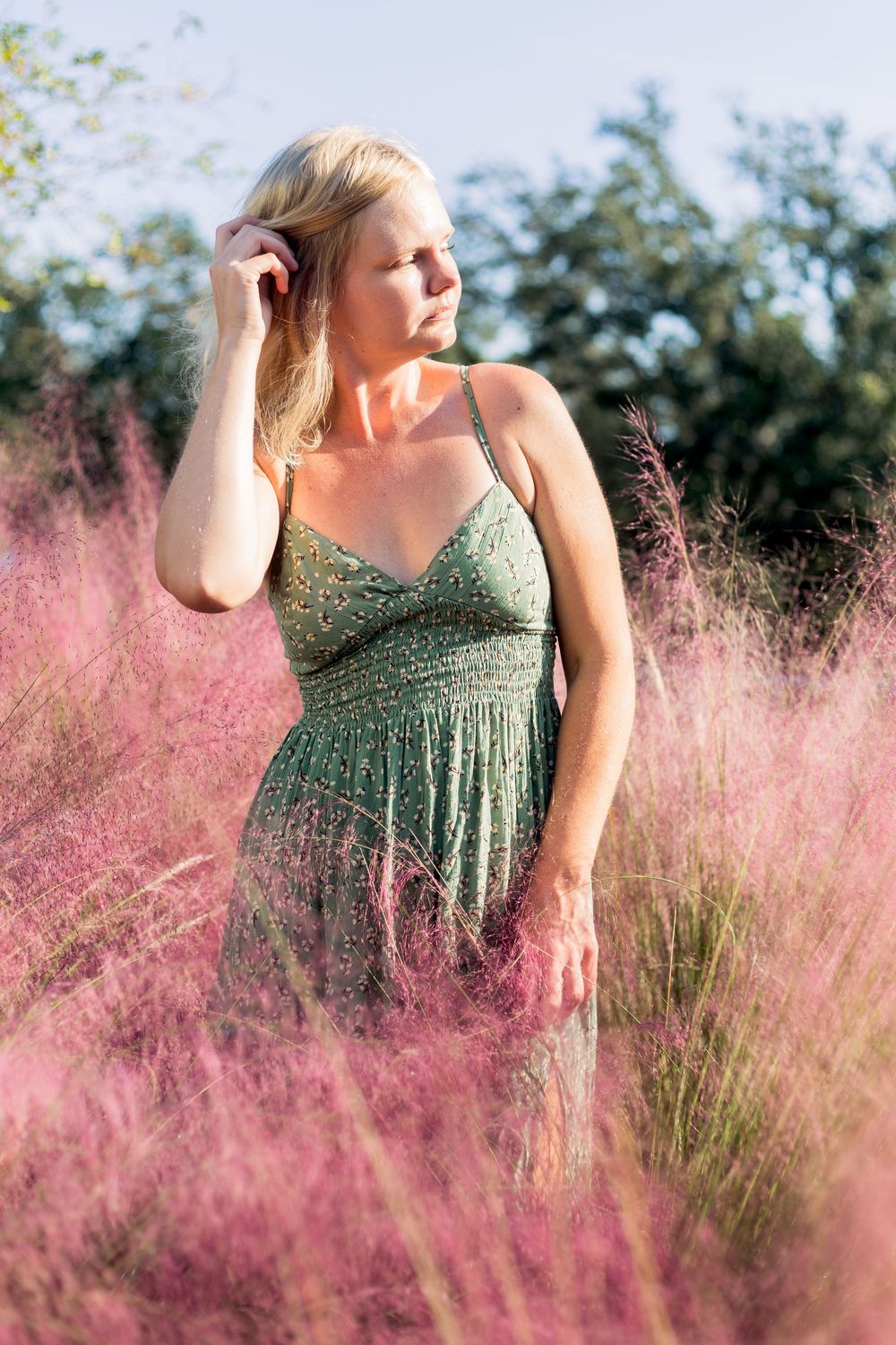 A person in a green dress stands thoughtfully among pink muhly grass on a sunny day.