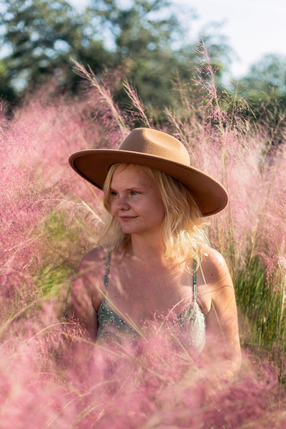 A person in a wide-brimmed brown hat sits among pink muhly grass in dreamy golden light.