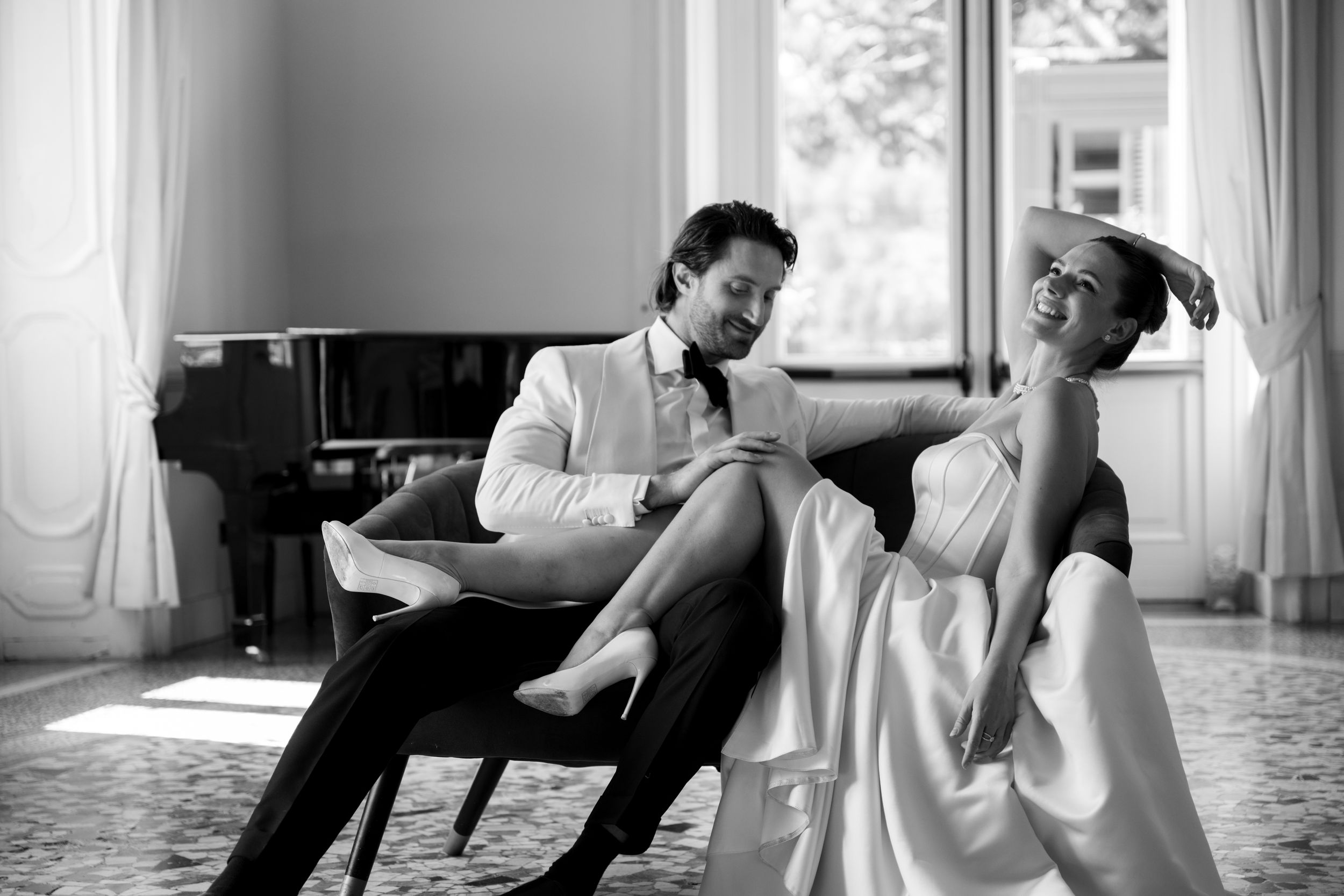 Two people lounge casually in a bright living room with a piano, captured in an intimate black and white moment.