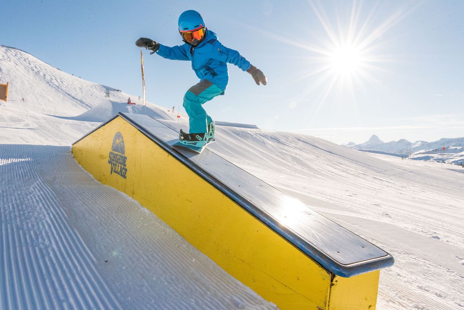 Snowboarder in bright blue gear sliding along a yellow and black rail against snowy mountain backdrop and bright sun.