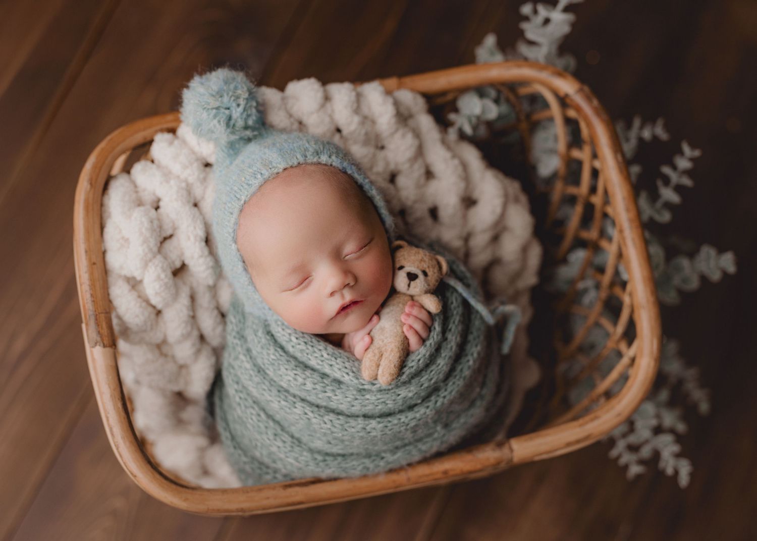 A sleeping baby swaddled in mint green fabric rests peacefully in a woven basket with cozy white knit blanket. Studio newborn photography
