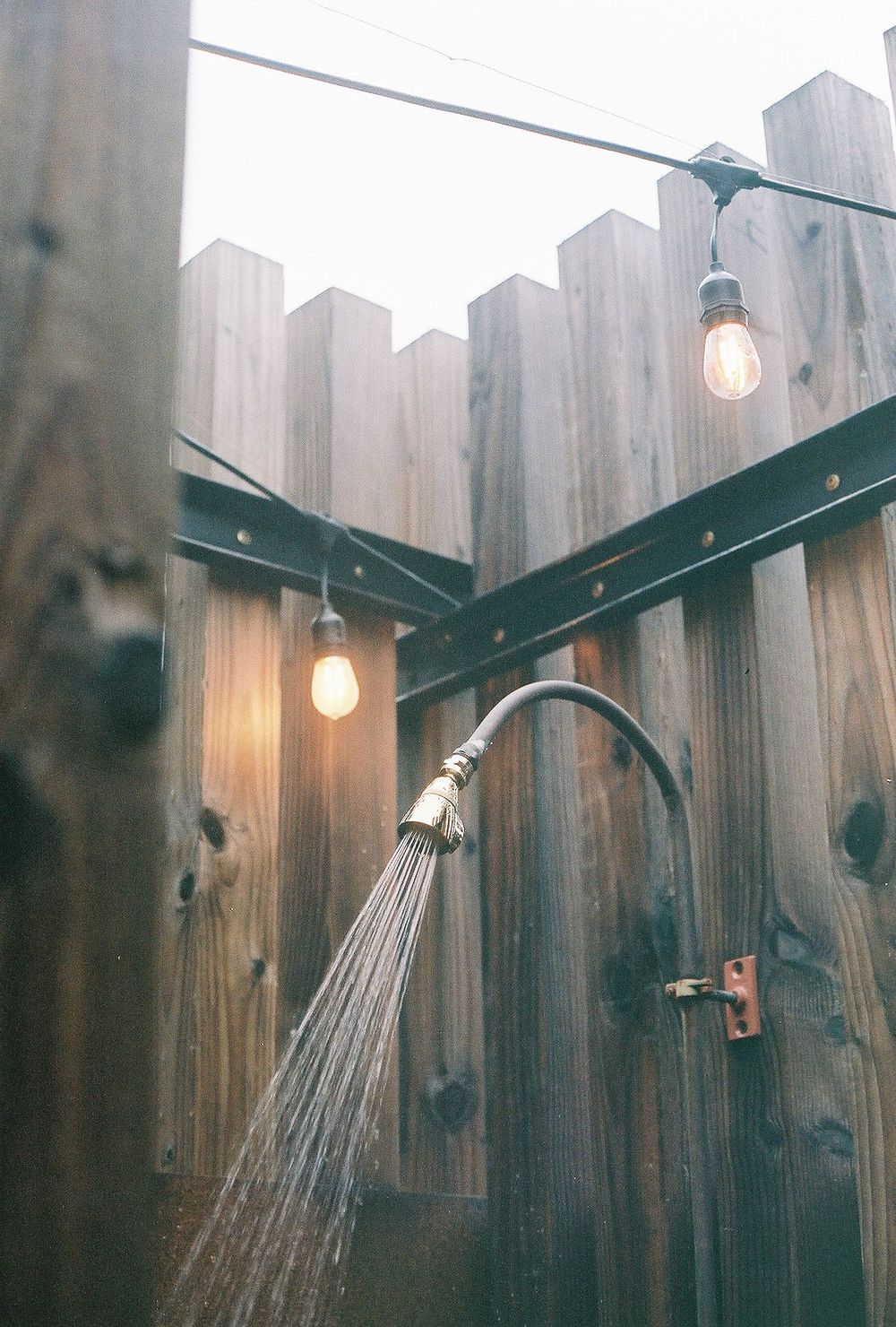Outdoor shower head mounted on wooden fence with string lights creating ambient lighting.