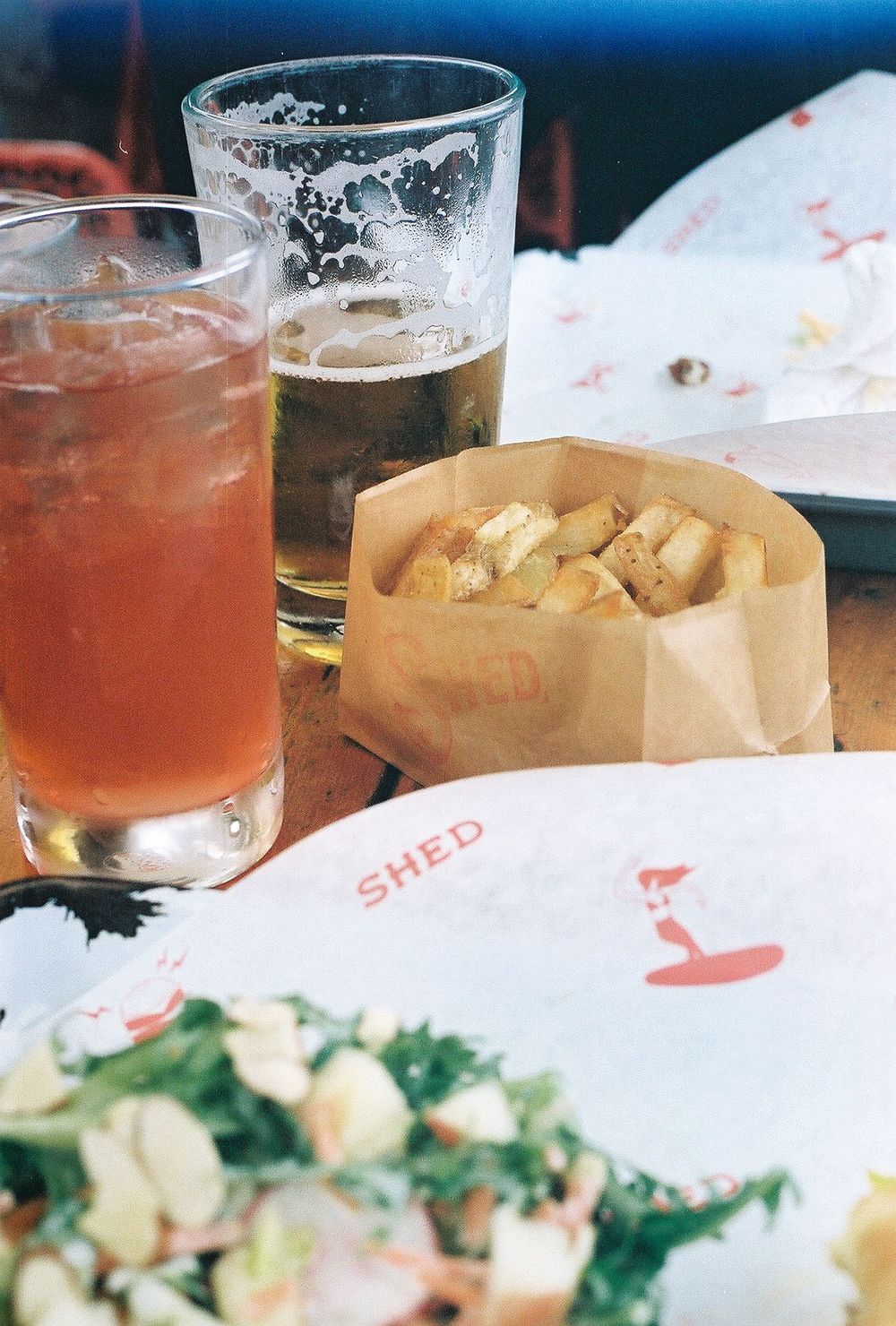 Two drinks and a brown paper bag of snacks on a restaurant table with plates and food.