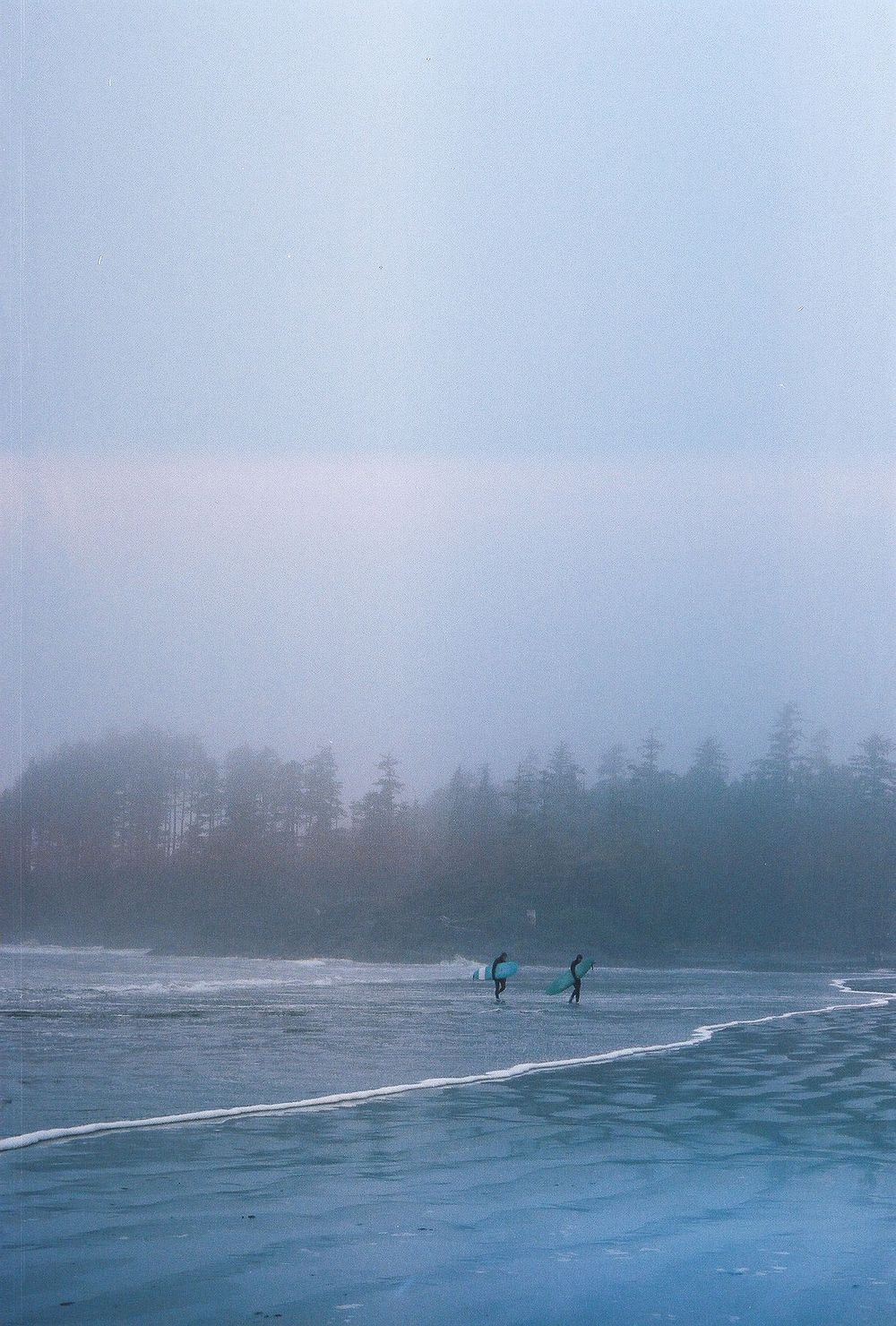 Distant figures surfing on misty ocean waves near a forested coastline.