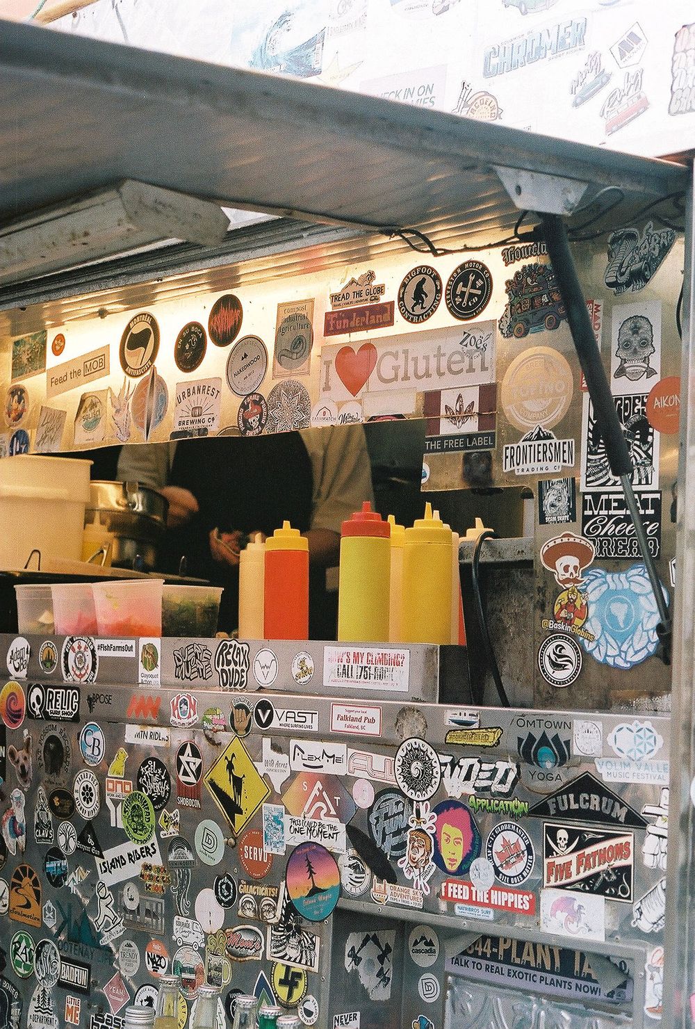 A food cart decorated with stickers and condiment bottles displays various sauce options and gluten-free signage.