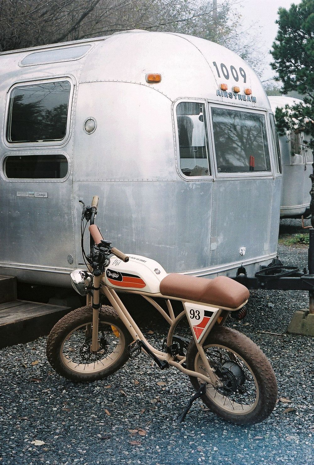 A vintage motorcycle parked next to a silver Airstream trailer in a gravel lot.
