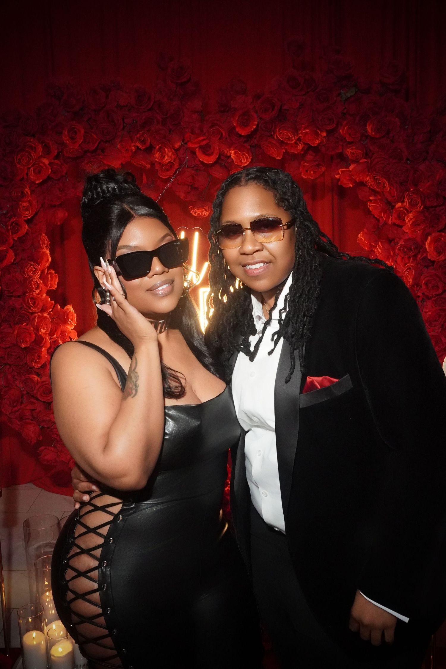 Couple in formal black attire poses against red rose wall at elegant event.