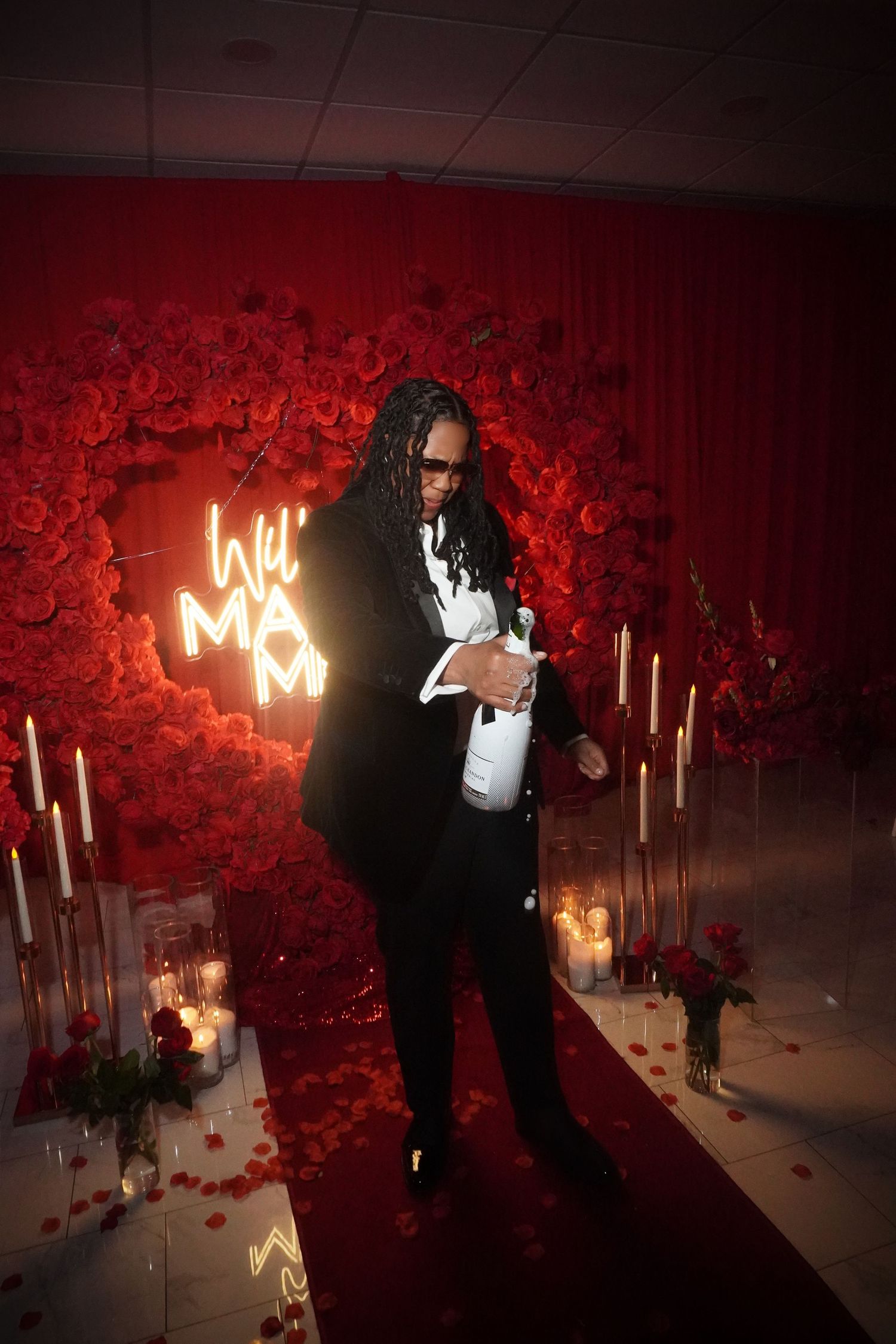 A stylish figure poses with champagne by a neon 'Will You Marry Me' sign surrounded by red roses.