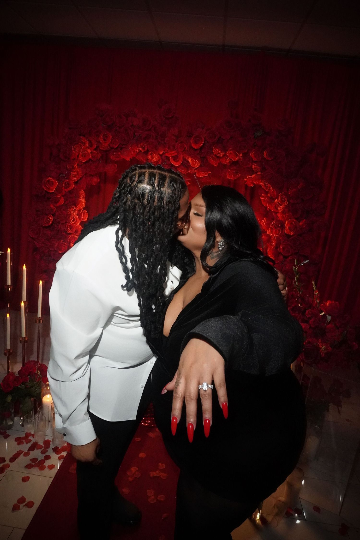 Couple shares intimate moment against dramatic red rose wall with glowing candles.