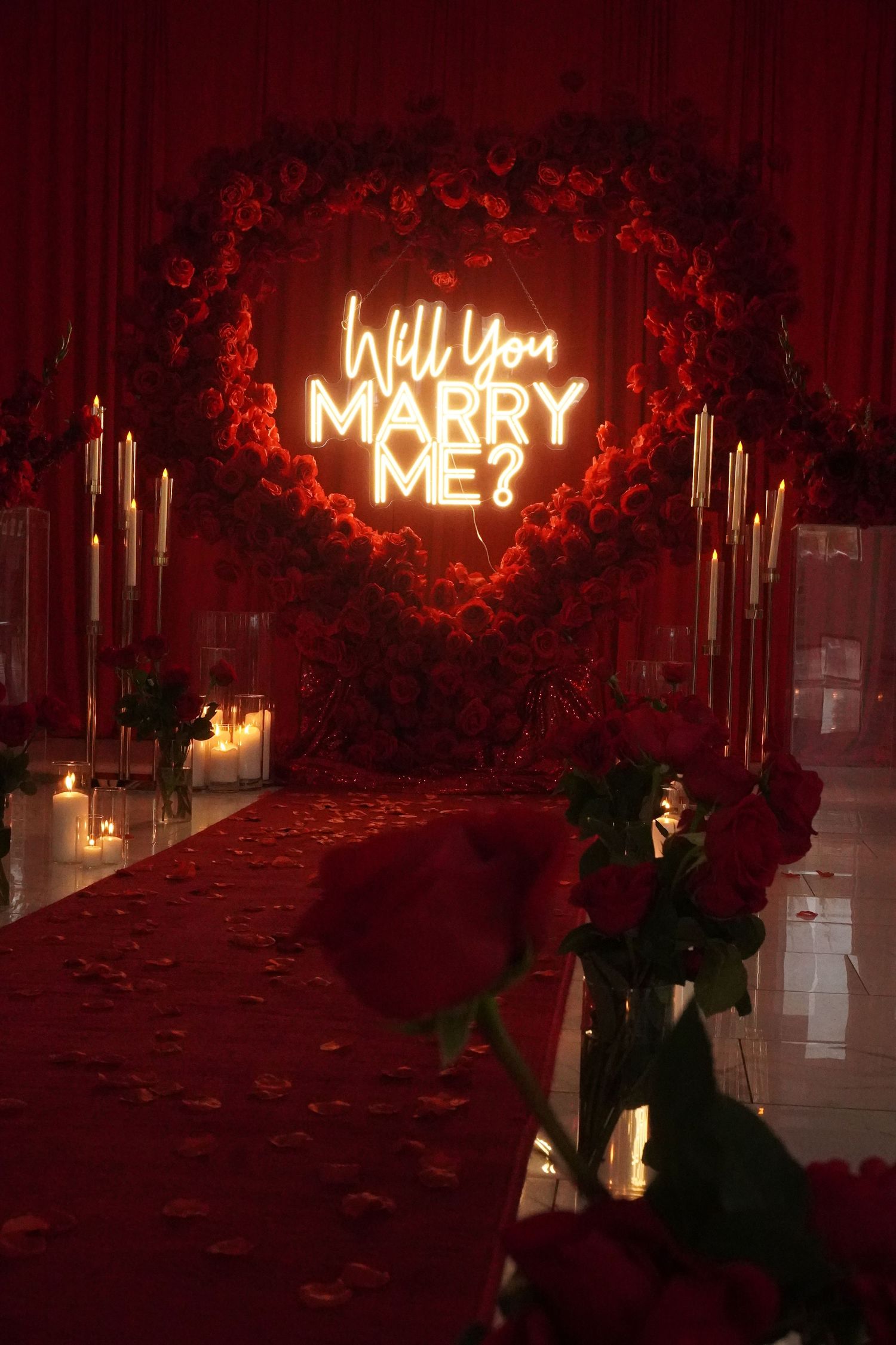 A romantic proposal setup with candles, red roses, and a glowing 'Will You Marry Me' neon sign.