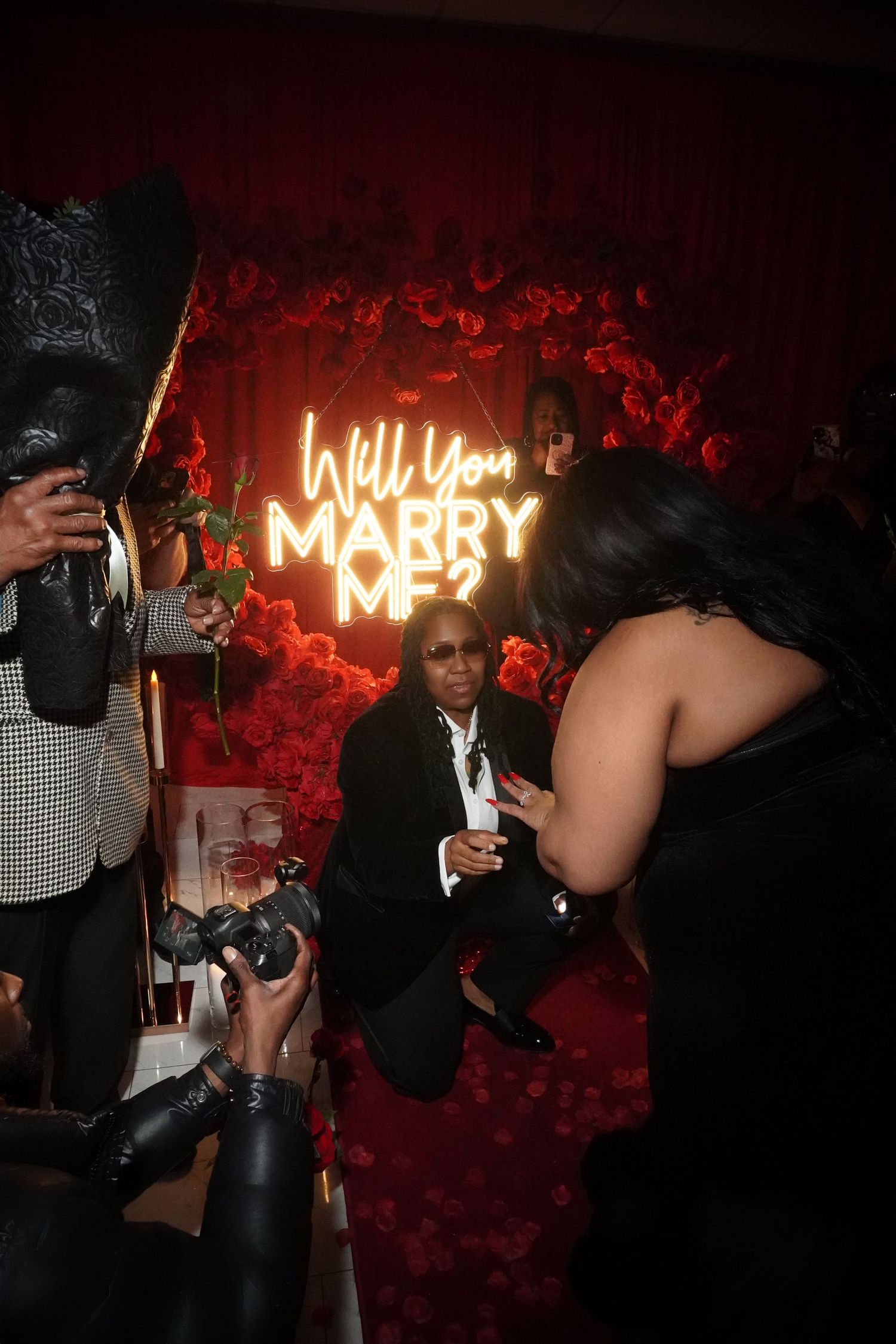 Marriage proposal scene with neon sign and photographers capturing the moment against red rose backdrop.
