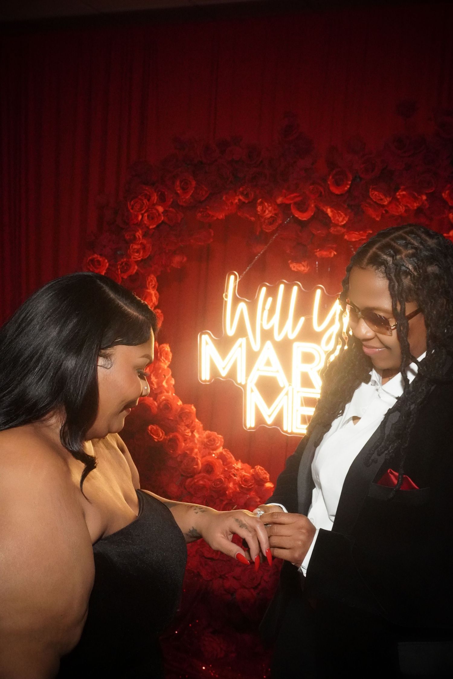 Couple shares romantic proposal moment in front of neon 'Will You Marry Me' sign with red balloon backdrop.