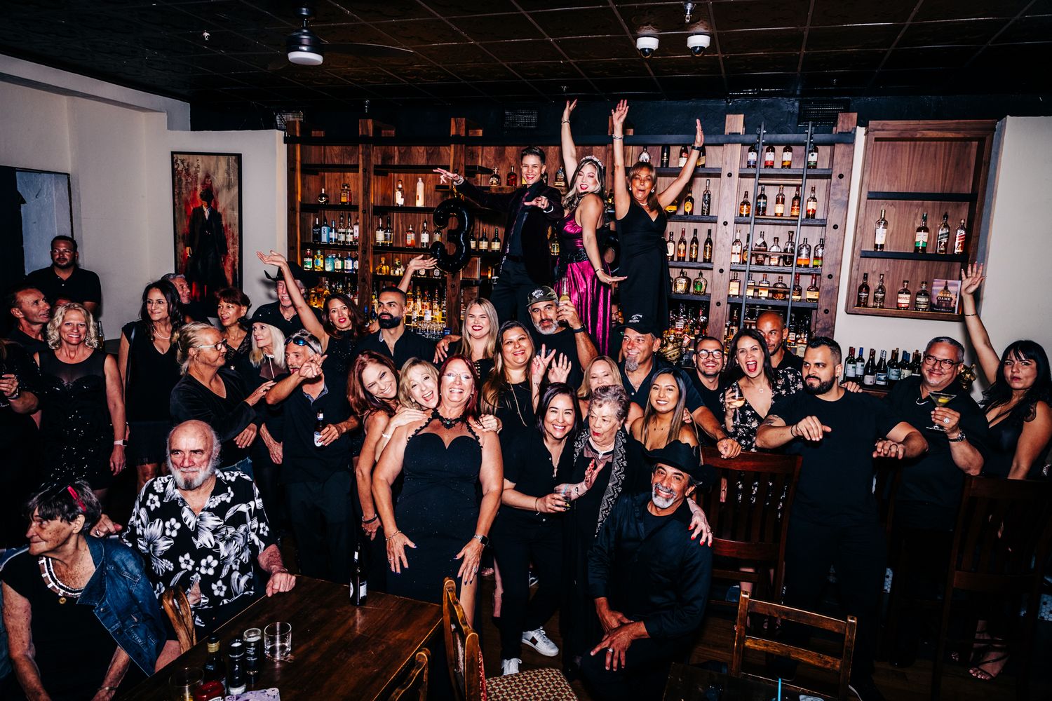 Large group of energetic people celebrating at a dimly lit bar with exposed brick walls and shelves of bottles at an event in lake worth.