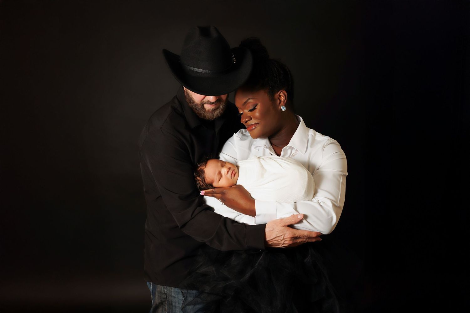 Parents in black cowboy hats tenderly hold their newborn baby against dark studio backdrop.