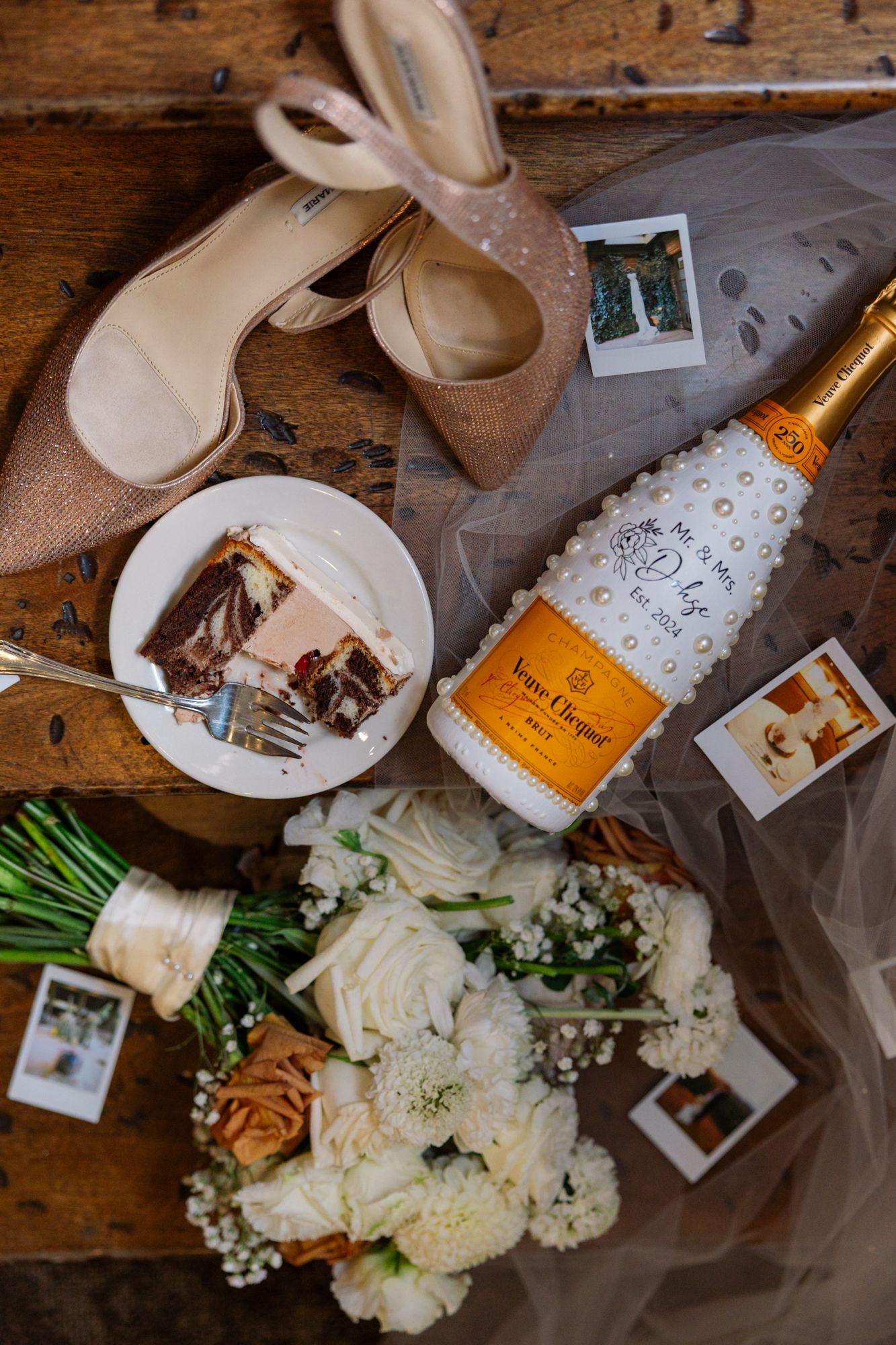 Wedding details including champagne, beige heels, white floral bouquet, cake slice and polaroid photos on wooden surface.