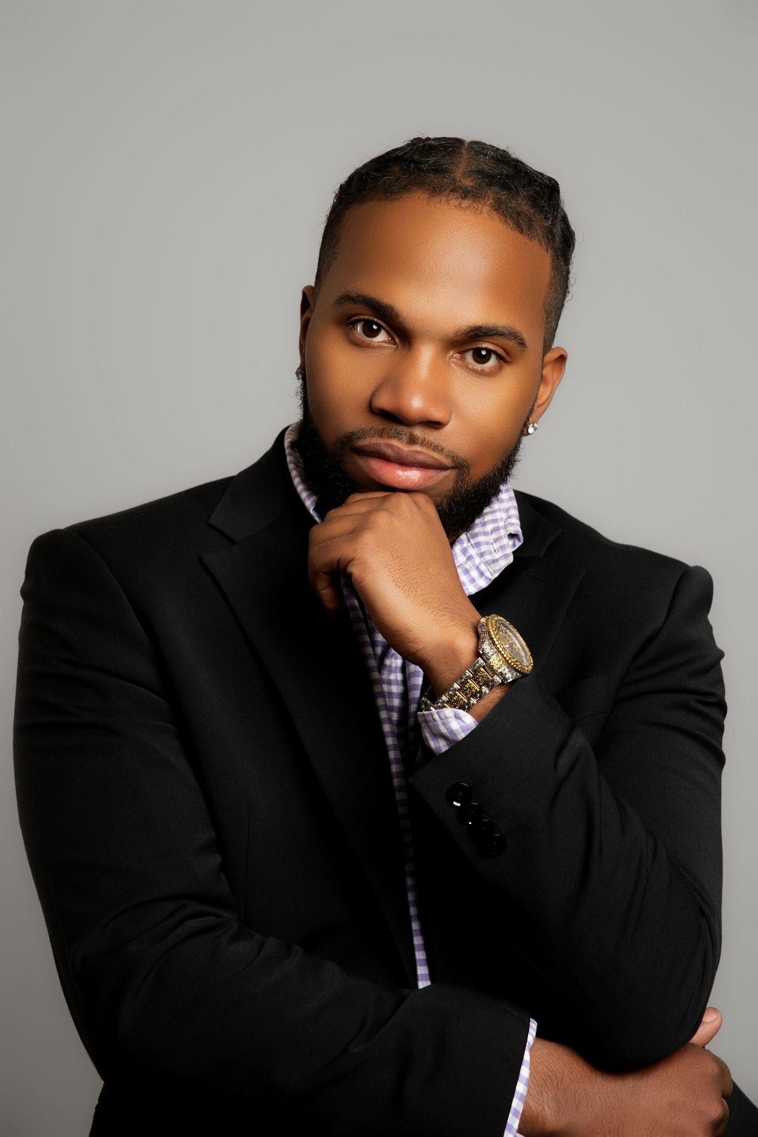 Professional headshot portrait in black suit and checkered shirt with thoughtful pose against gray background.