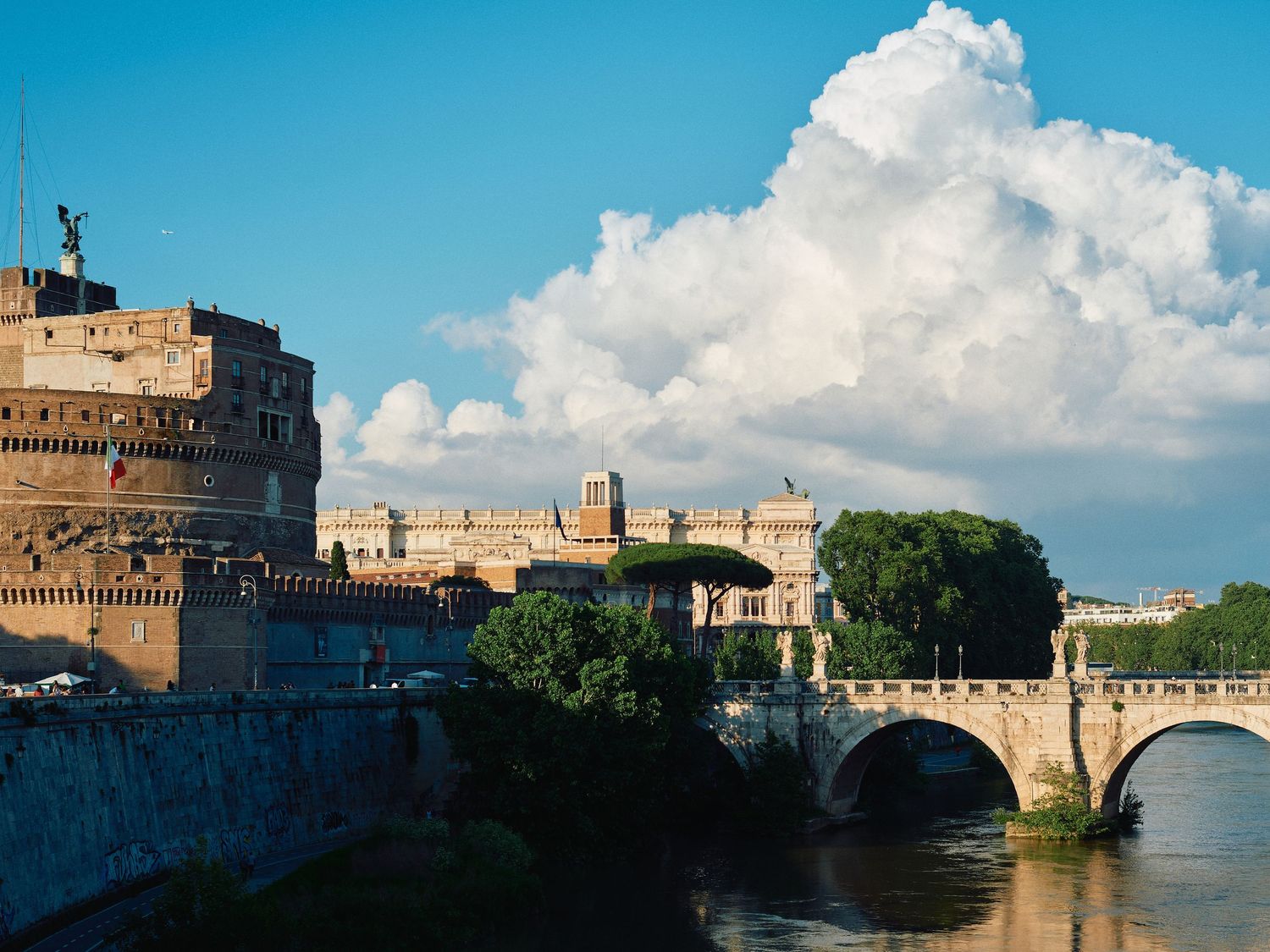 View of Castel Sant'Angelo and Sant'Angelo Bridge over the Tiber River in Rome with puffy white clouds in blue sky.