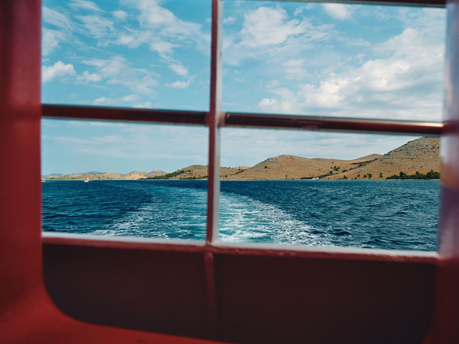 View through a red boat window showing blue ocean waves and rolling coastal hills under a cloudy sky.