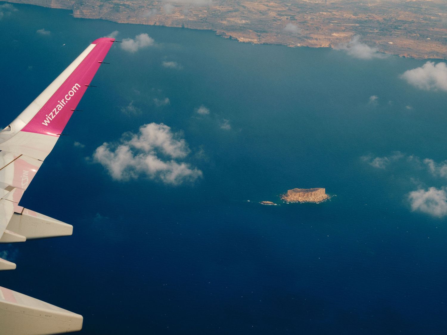 Aerial view of pink airplane wing over deep blue ocean water with small island visible below through scattered clouds.