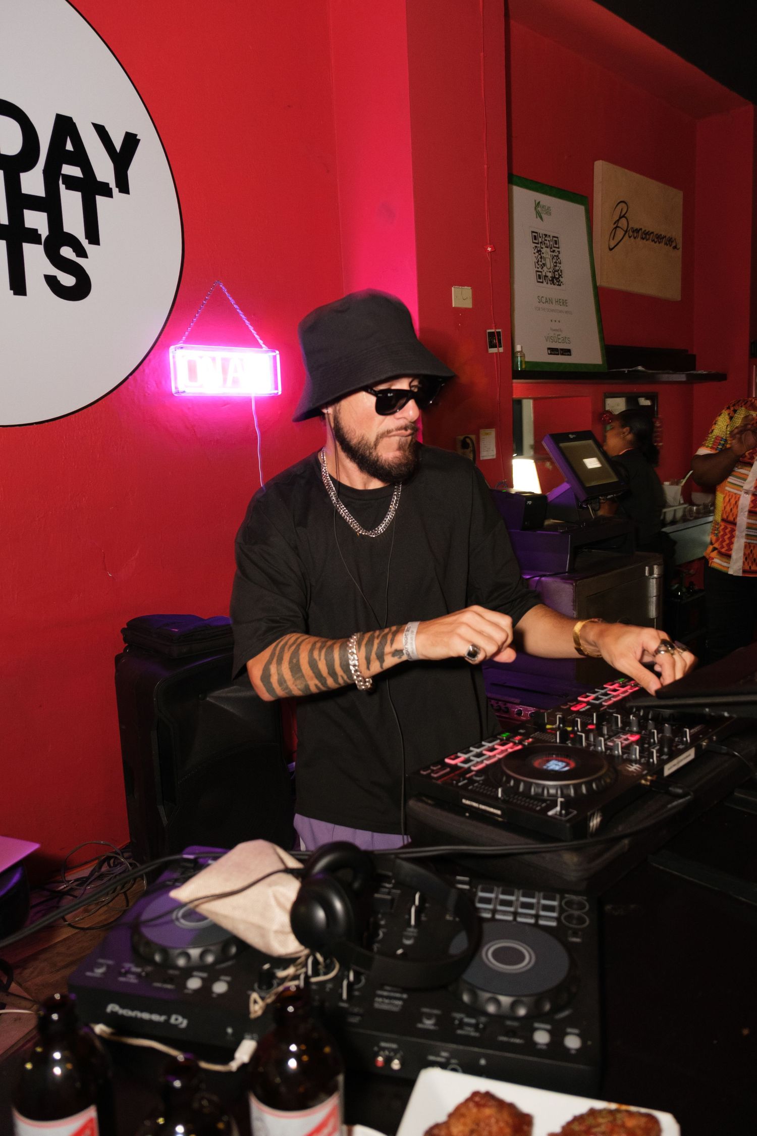 DJ in black attire working at a mixer setup with Pioneer equipment in a red-lit nightclub venue with neon signage.